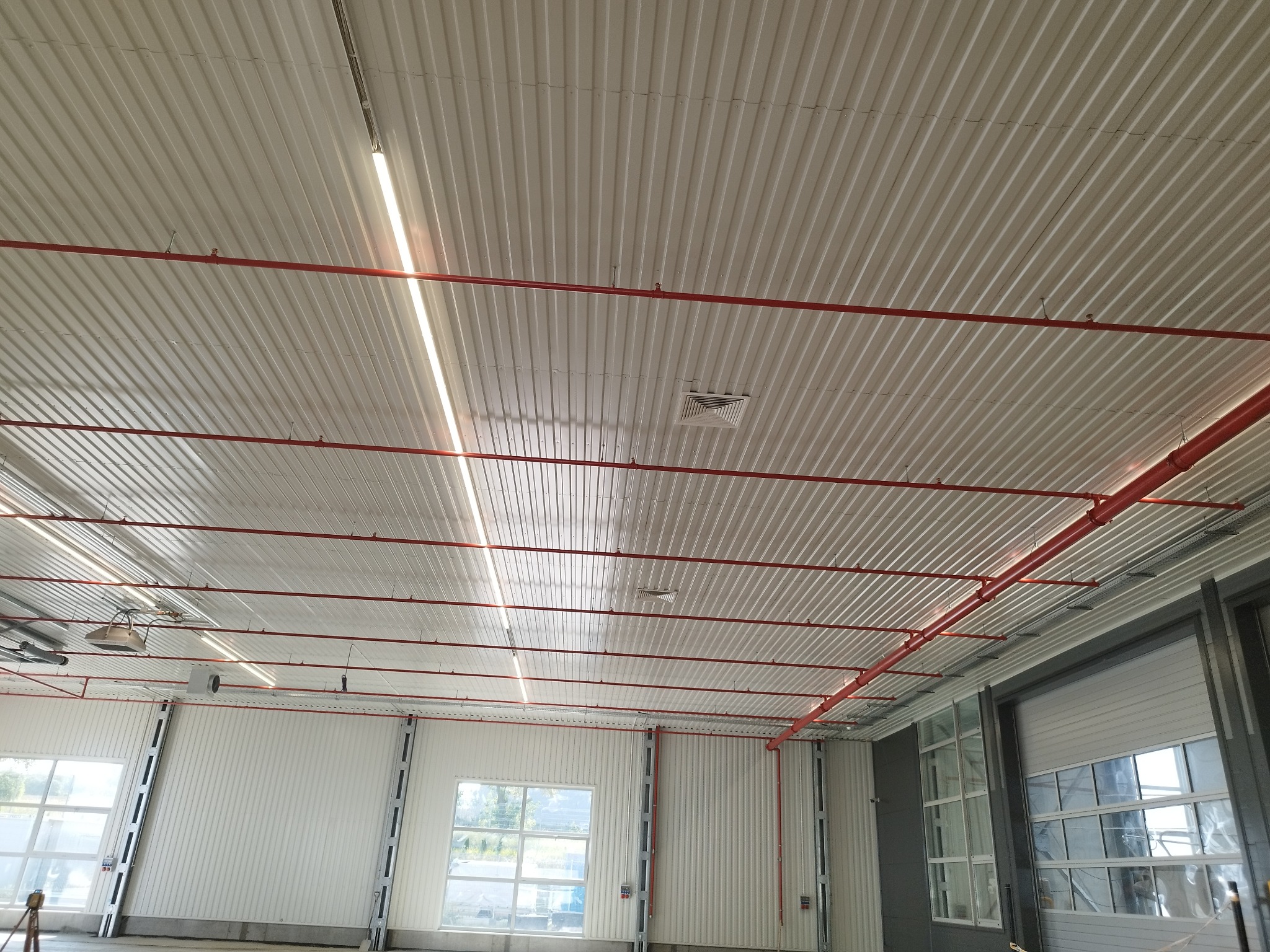 Wnętrze hali przemysłowej z widoczną instalacją przeciwpożarową (czerwone rury) pod sufitem z blachy trapezowej i oświetleniem liniowym LED.