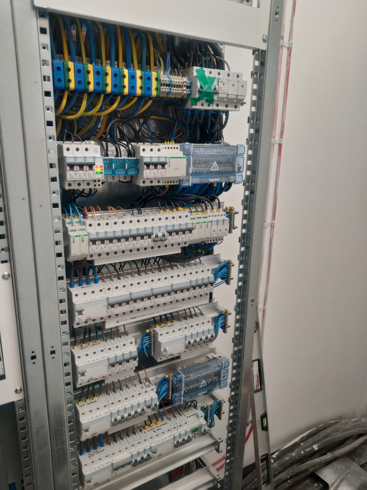 Szczegółowe ujęcie rozdzielnicy elektrycznej z licznymi zabezpieczeniami i przewodami oznaczonymi kolorami, zamontowanej w metalowej obudowie z poziomnicą opartą o ścianę.