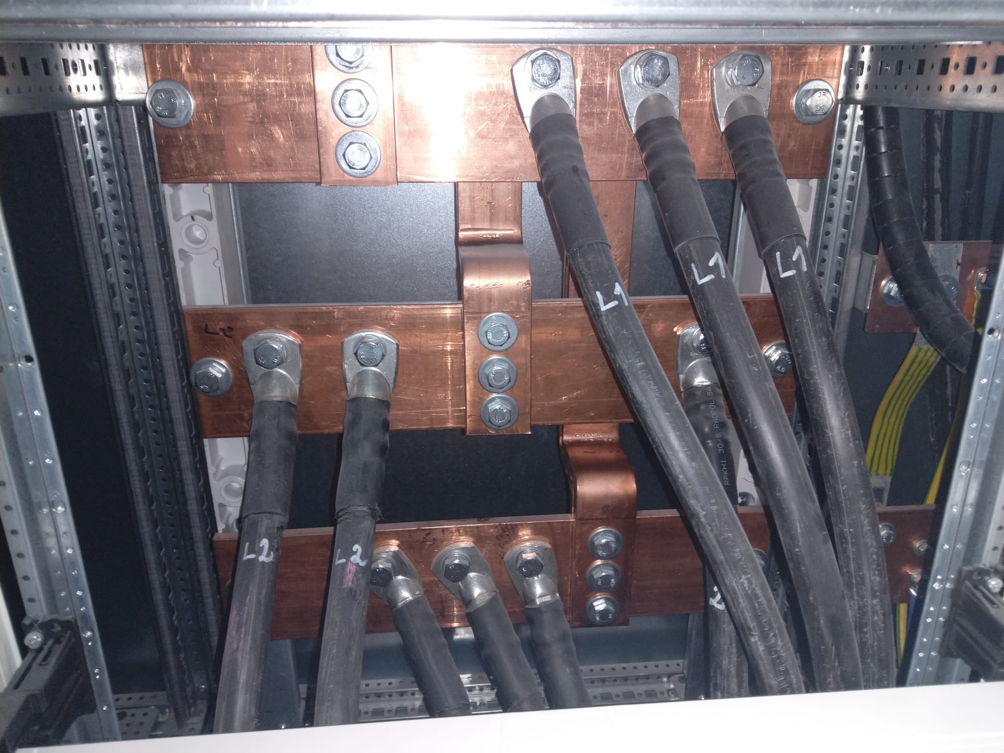 Miedziane szyny z podłączonymi grubymi przewodami oznaczonymi L1 i L2 w rozdzielnicy elektrycznej, widoczne śruby mocujące i elementy konstrukcyjne