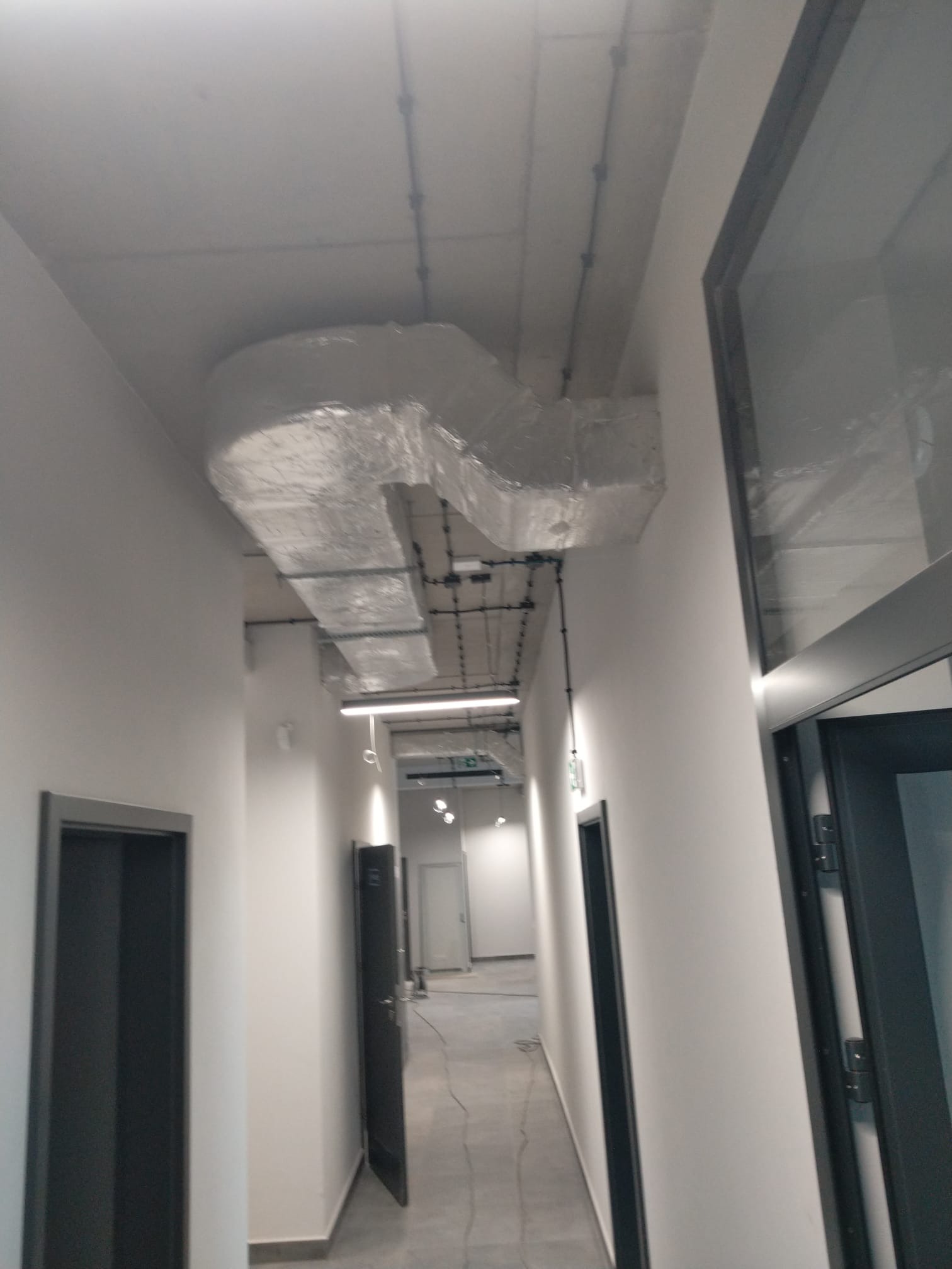 Korytarz biurowy z widoczną instalacją wentylacyjną pod sufitem, zabezpieczoną folią ochronną, oraz przewodami elektrycznymi.