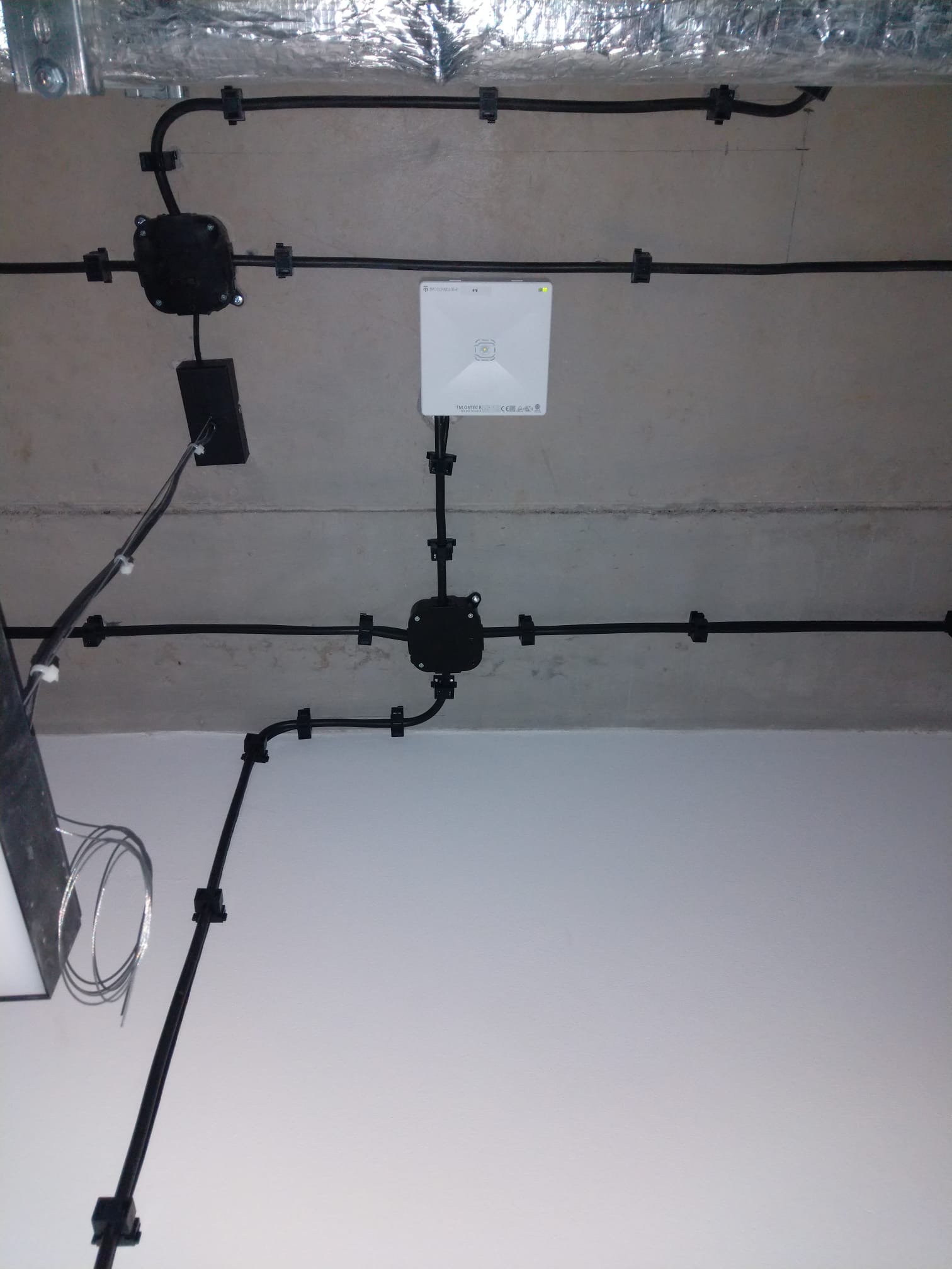 Czarna instalacja kablowa przymocowana do sufitu, z widoczną puszką rozgałęźną, punktem dostępowym Wi-Fi i fragmentem wentylacji.