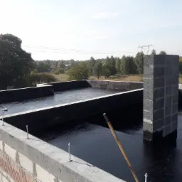 Prace dekarskie na płaskim dachu: robotnik rozprowadza czarną masę bitumiczną na betonowej powierzchni, obok komin z szarego betonu komórkowego i wystające metalowe śruby mocujące na obrzeżach...