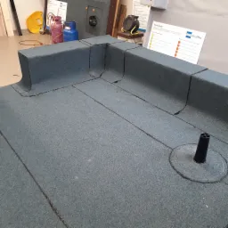 Model płaskiego dachu pokrytego papą z widocznymi zgrzewami, kominkiem wentylacyjnym i detalami obróbek blacharskich, prezentowany na ekspozycji z akcesoriami dekarskimi w tle.