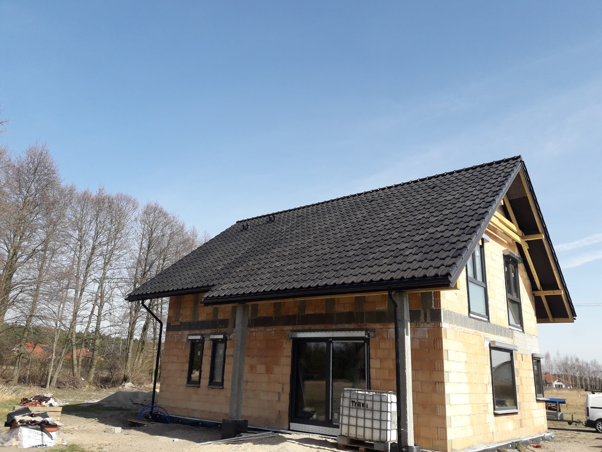 Dom w trakcie budowy z czarnym dachem z dachówki ceramicznej, widoczne ściany z pustaków ceramicznych, okna z czarnymi ramami i drewniana konstrukcja szczytu dachu, na zewnątrz paleta z pojemnikiem...