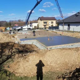 Wylewanie fundamentów domu jednorodzinnego z użyciem pompy do betonu; widoczny zbrojony szkielet fundamentu, pracownicy w odblaskowych kamizelkach i cień fotografa na pierwszym planie.