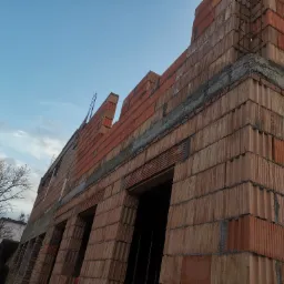 Surowa ściana zewnętrzna budynku w trakcie budowy, wykonana z czerwonej cegły ceramicznej, z widocznymi otworami okiennymi i elementami zbrojenia, na tle błękitnego nieba i drzewa bez liści.