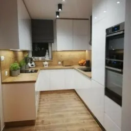 Nowoczesna kuchnia z białymi szafkami, drewnianym blatem i podłogą imitującą drewno, oświetlona punktowymi lampami sufitowymi i z roletą w oknie.