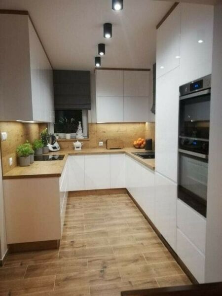 Nowoczesna kuchnia z białymi szafkami, drewnianym blatem i podłogą imitującą drewno, oświetlona punktowymi lampami sufitowymi i z roletą w oknie.