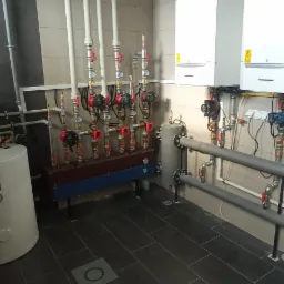 Węzeł rozdzielczy ogrzewania podłogowego z dwoma piecami CO, widoczne rury, zawory, pompy obiegowe, grzejniki oraz odpływ podłogowy.