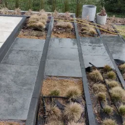 Fragment ogrodu z ułożonymi jasnymi płytami tarasowymi, ciemnoszarymi płytami ścieżki i rabatami z ozdobną trawą, widoczny system nawadniania i żółty wąż ogrodowy.