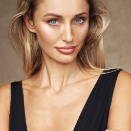 Aneta Byczyk - Portret kobiety w czarnym stroju z delikatnym uśmiechem, podkreślonym makijażem i blond włosami w studio z neutralnym tłem.