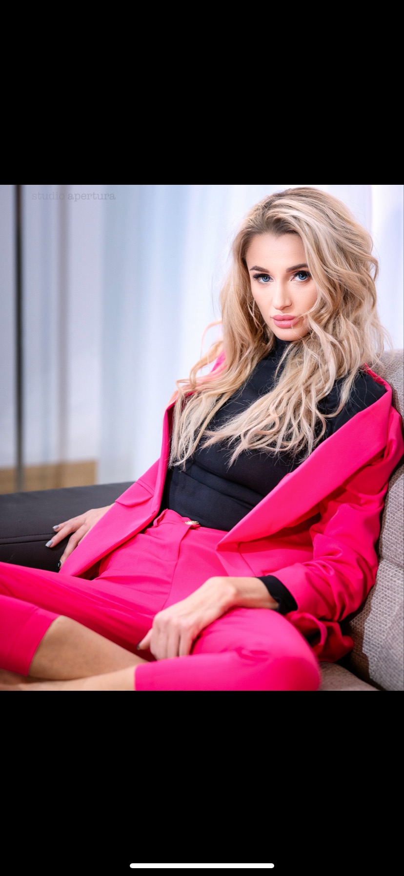 Kobieta w różowym garniturze i czarnym topie, siedząca na kanapie, z delikatnym uśmiechem i blond włosami, sfotografowana w studio.