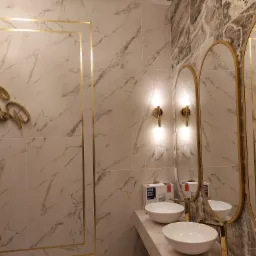 Elegancka łazienka z marmurowymi płytkami, złotymi ramami luster i dekoracyjnym napisem na ścianie, oświetlona kinkietami.