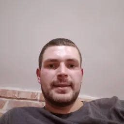 Portret mężczyzny z krótką brodą i włosami, ubrany w szarą koszulkę, z fragmentem ceglanej ściany w tle.