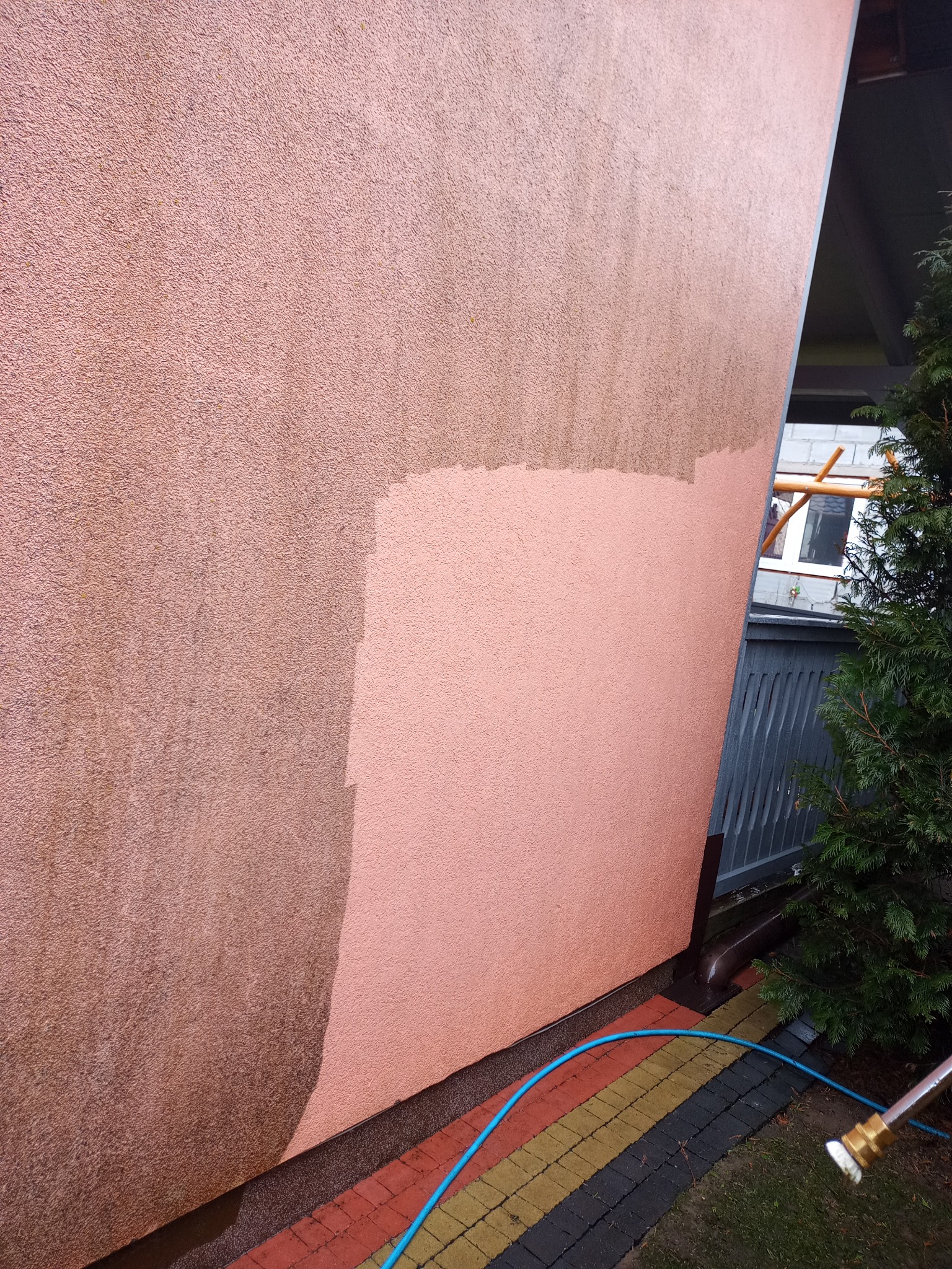 Częściowo wyczyszczona, brudna elewacja domu o różowej barwie, widoczny efekt przed i po, niebieski wąż leżący na kostce brukowej.