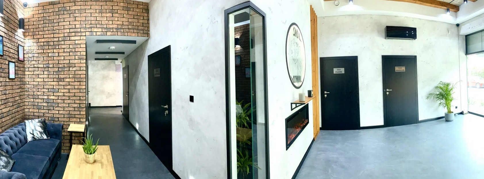Wnętrze szkoły językowej z ceglaną ścianą, niebieską sofą i przeszklonym pomieszczeniem z roślinami, widoczne drzwi z oznaczeniami sal.