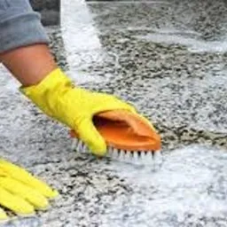 Osoba w żółtych rękawicach szoruje kamienną powierzchnię szczotką, widoczna piana i ślady czyszczenia.