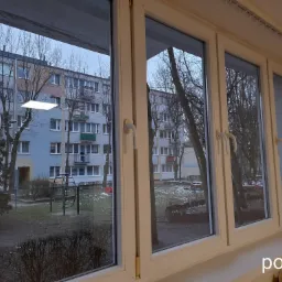 Widok z okna na blok mieszkalny i plac zabaw w pochmurny dzień, trzy białe ramy okienne z widocznym napisem 'po' na parapecie.