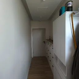 Wnętrze wąskiego korytarza z zabudową szaf z szufladami w kolorze białym, drewnianą podłogą i białą ścianą po lewej stronie, widoczne detektory dymu na suficie.