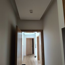 MW Bau Marcel Wende - Korytarz po remoncie z nową podwieszaną płytą kartonowo-gipsową na suficie, widok z dołu na białe ściany, drewniane ościeżnice i panele podłogowe. Czarny kubeł stoi w rogu.
