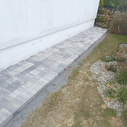 MW Bau Marcel Wende - Ułożony chodnik z szarej kostki brukowej prowadzący wzdłuż białego budynku w kierunku czarnej bramki ogrodzeniowej, oddzielony od trawnika pasem drobnego żwiru.