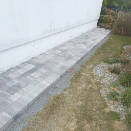 Ułożony chodnik z szarej kostki brukowej prowadzący wzdłuż białego budynku w kierunku czarnej bramki ogrodzeniowej, oddzielony od trawnika pasem drobnego żwiru.