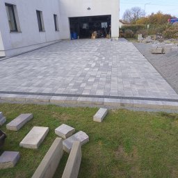 MW Bau Marcel Wende - Ułożona kostka brukowa na podjeździe przed garażem domu jednorodzinnego, z widocznymi niedokończonymi obrzeżami i stertą kostki na trawniku.