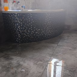 MW Bau Marcel Wende - Szara łazienka z wanną obłożoną mozaiką, butelka z napojem na rancie wanny, widoczne płytki podłogowe z efektem betonu.