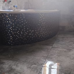 MW Bau Marcel Wende - Zaokrąglona obudowa wanny wykończona mozaiką w odcieniach szarości i czerni, dopasowana do szarej, błyszczącej podłogi z płytek w łazience, widoczne baterie i fragment wanny.