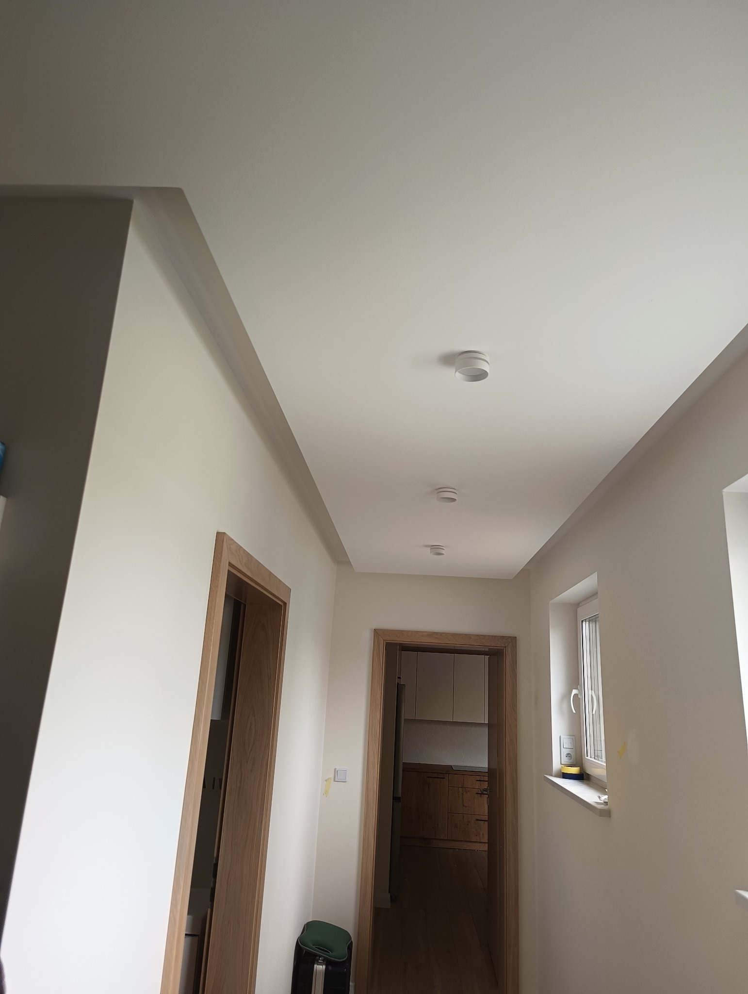 Korytarz w jasnych barwach po remoncie, z widocznymi trzema okrągłymi lampami sufitowymi, drewnianymi ościeżnicami drzwi i widokiem na kuchnię w tle.