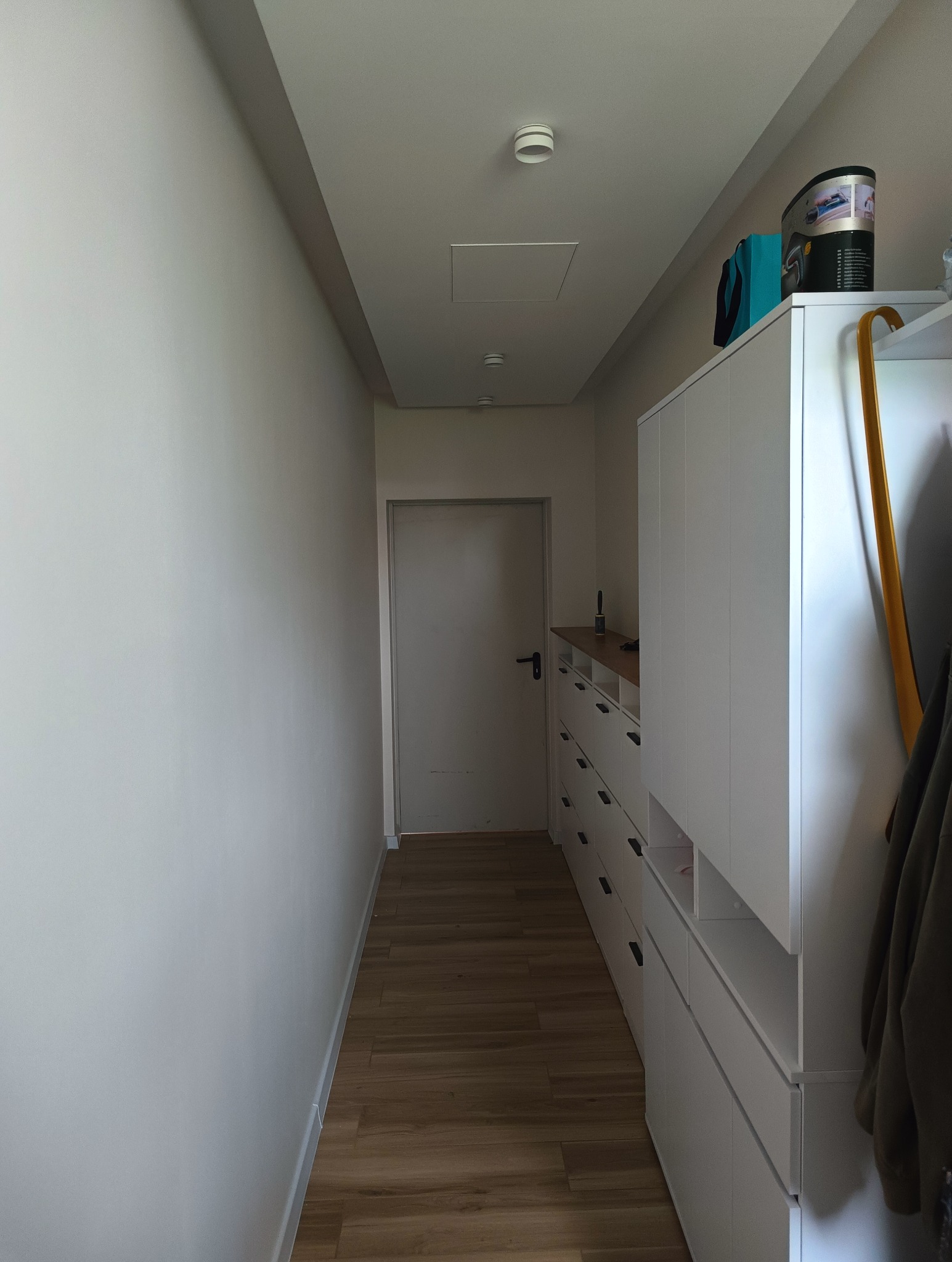 Wnętrze wąskiego korytarza z zabudową szaf z szufladami w kolorze białym, drewnianą podłogą i białą ścianą po lewej stronie, widoczne detektory dymu na suficie.