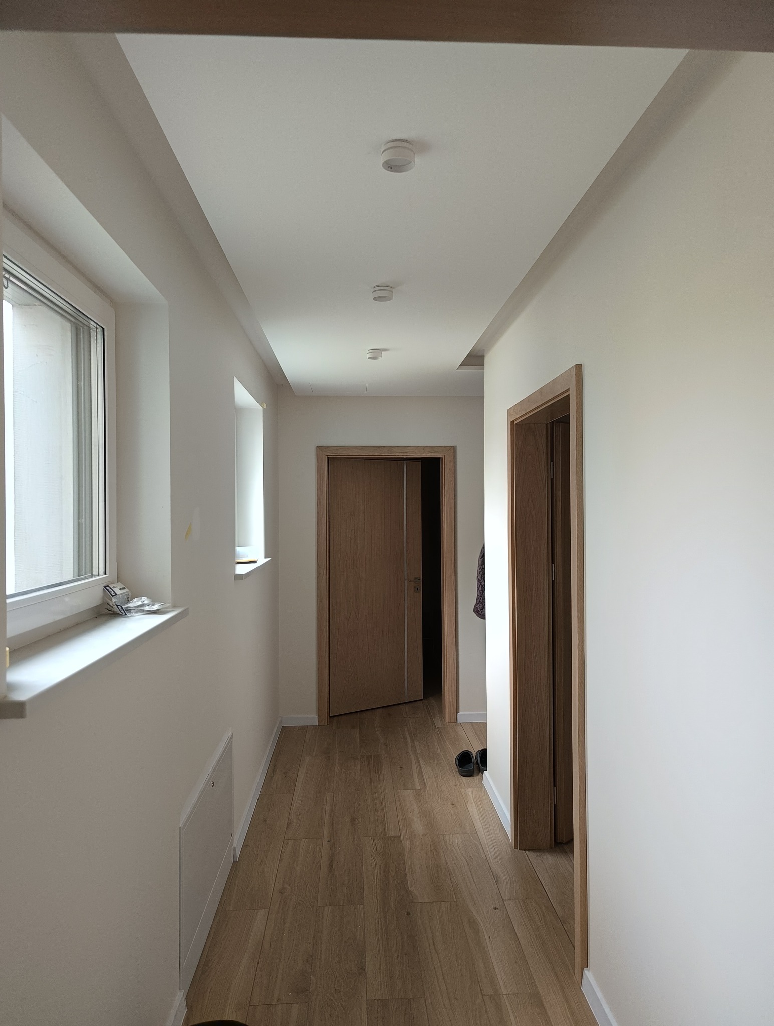 Korytarz w jasnych barwach z drewnopodobną podłogą, białymi ścianami i dwiema parami drzwi z drewnianymi ościeżnicami, widok na sufit z oświetleniem punktowym.