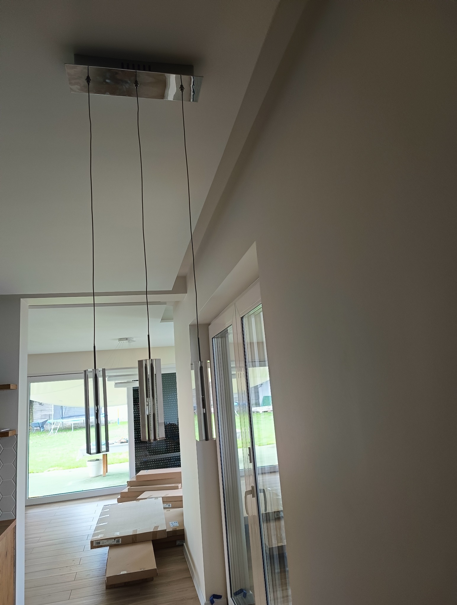 Nowoczesna, chromowana lampa wisząca z trzema podłużnymi kloszami, zamontowana w nowym domu podczas wykańczania wnętrz, z widocznymi kartonami na podłodze.