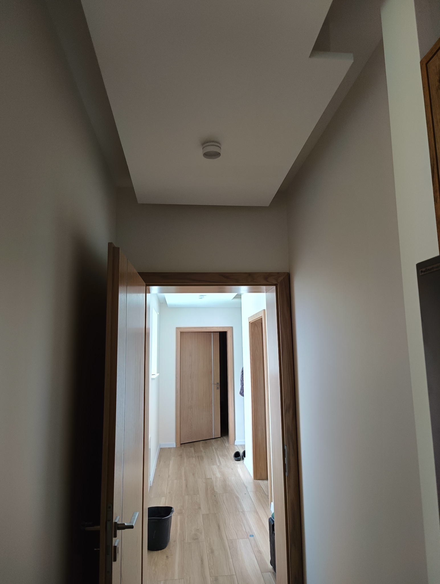 Korytarz po remoncie z nową podwieszaną płytą kartonowo-gipsową na suficie, widok z dołu na białe ściany, drewniane ościeżnice i panele podłogowe. Czarny kubeł stoi w rogu.