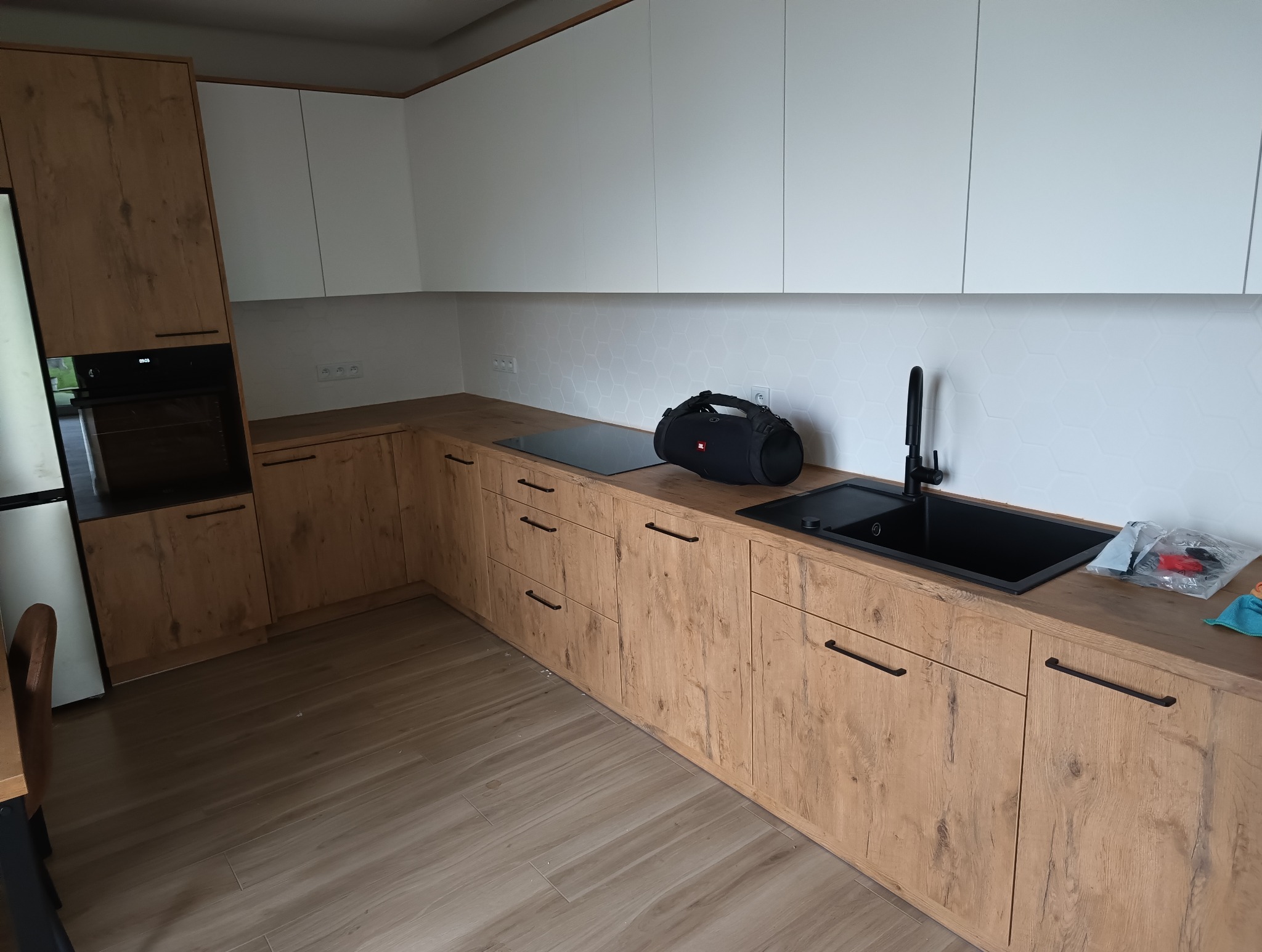 Nowoczesna kuchnia z drewnopodobnymi szafkami dolnymi, białymi szafkami górnymi i heksagonalnymi płytkami na ścianie, czarny zlew i bateria, płyta indukcyjna, na blacie leży głośnik JBL i akcesoria...