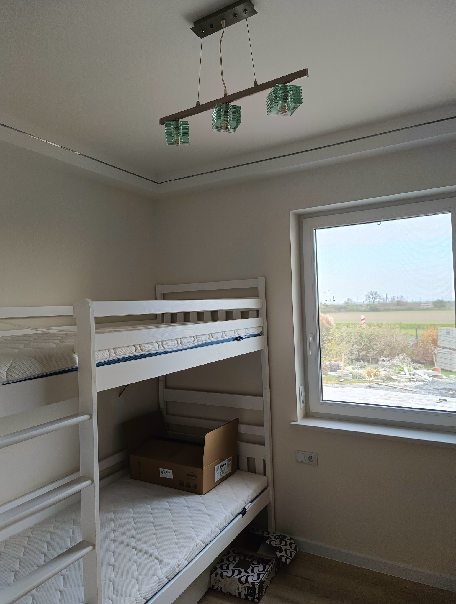 Pokój dziecięcy z białym łóżkiem piętrowym, kartonem na materacu i nowoczesną lampą sufitową z zielonymi kloszami, widok na zewnątrz przez okno.