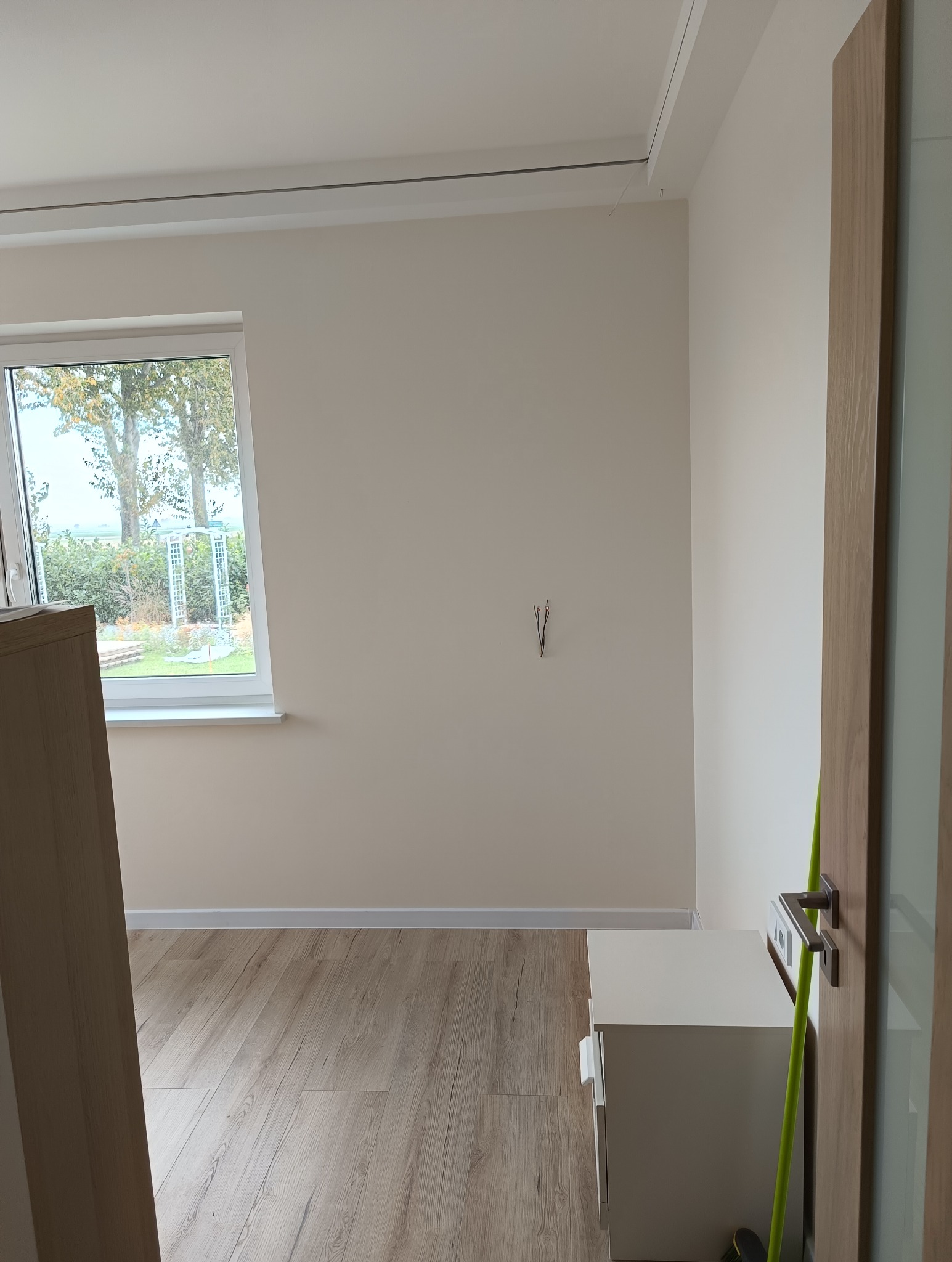 Pomalowane na beżowo wnętrze pokoju z drewnianą podłogą, białym oknem z widokiem na zieleń, fragmentem szafki i wystającymi przewodami elektrycznymi ze ściany.