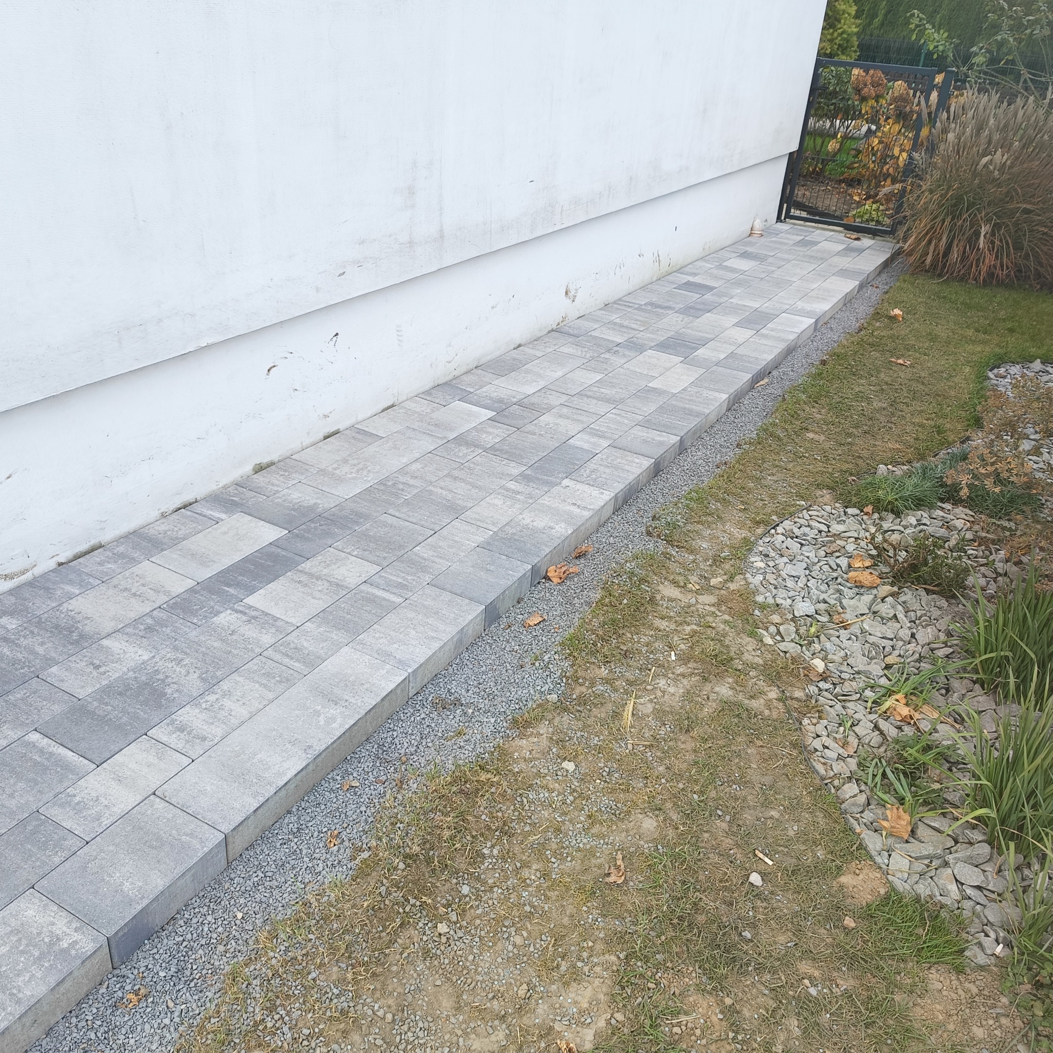 Ułożony chodnik z szarej kostki brukowej prowadzący wzdłuż białego budynku w kierunku czarnej bramki ogrodzeniowej, oddzielony od trawnika pasem drobnego żwiru.