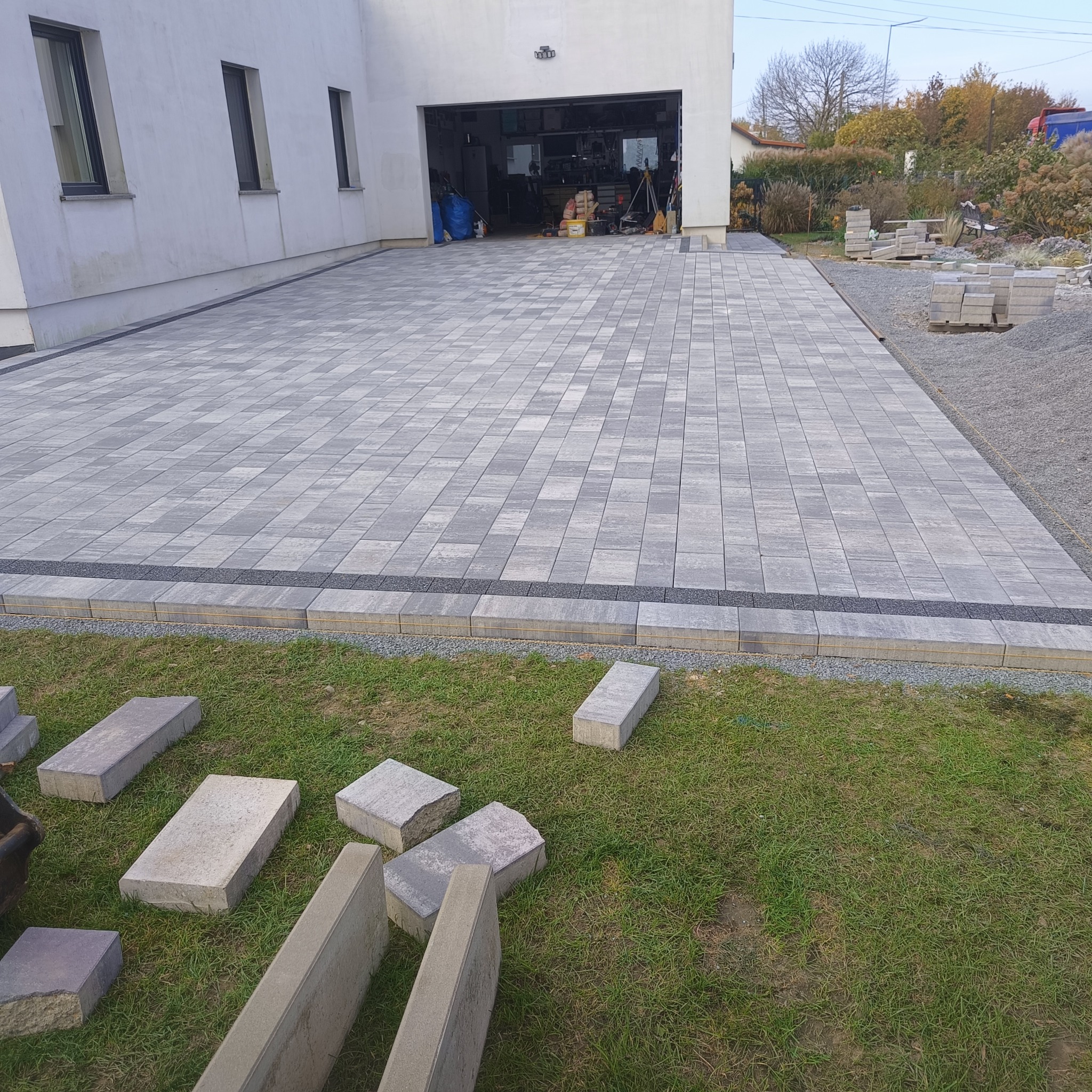 Ułożona kostka brukowa na podjeździe przed garażem domu jednorodzinnego, z widocznymi niedokończonymi obrzeżami i stertą kostki na trawniku.