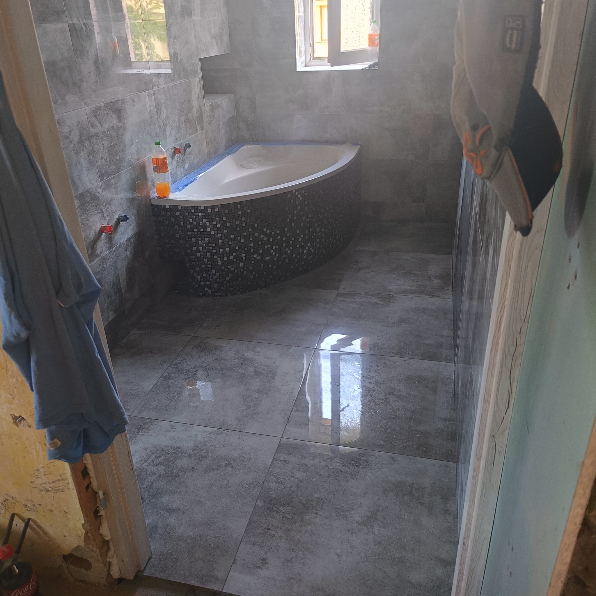 Szare płytki podłogowe i ścienne w łazience z mozaikową obudową wanny narożnej, widoczne narzędzia i niedokończone wykończenie przy futrynie drzwi.