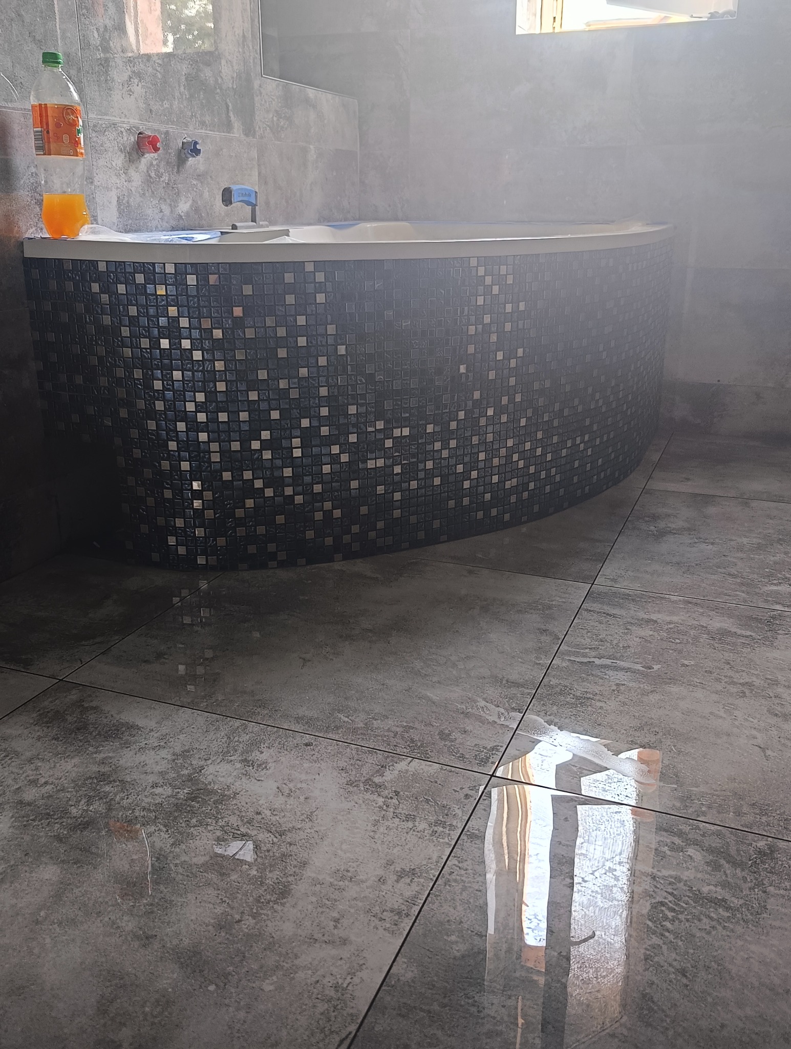 Szara łazienka z wanną obłożoną mozaiką, butelka z napojem na rancie wanny, widoczne płytki podłogowe z efektem betonu.