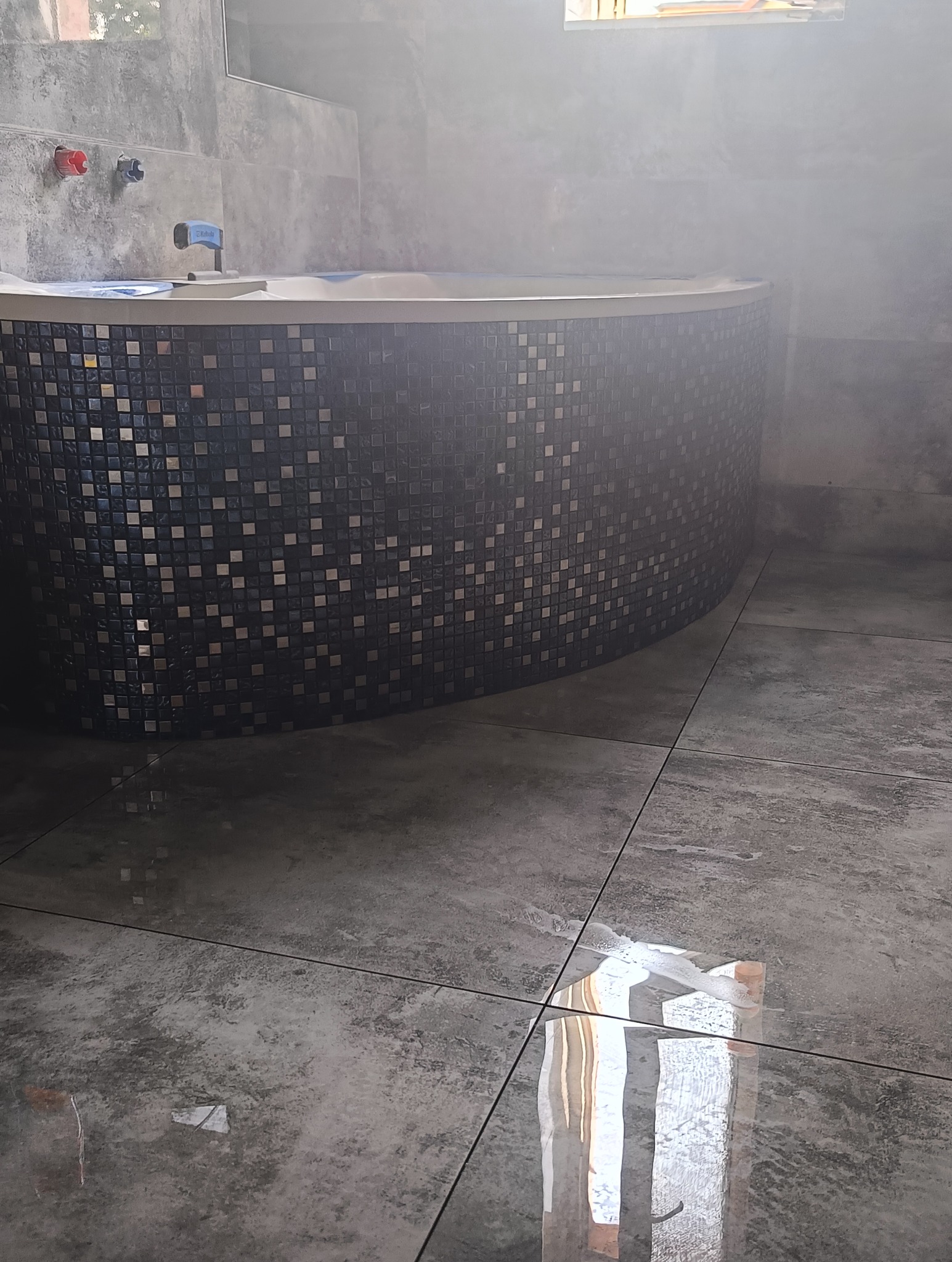 Zaokrąglona obudowa wanny wykończona mozaiką w odcieniach szarości i czerni, dopasowana do szarej, błyszczącej podłogi z płytek w łazience, widoczne baterie i fragment wanny.