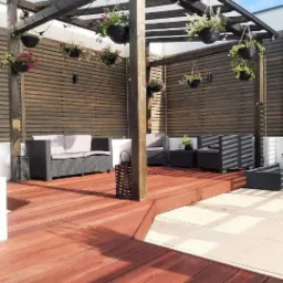 Zadaszony taras z drewnianą podłogą, rattanowymi meblami ogrodowymi i wiszącymi donicami z kwiatami, otoczony drewnianą ażurową ścianą.