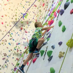 Chłopiec w zielonym stroju wspina się po ścianie wspinaczkowej z kolorowymi uchwytami, zabezpieczony uprzężą i linami.