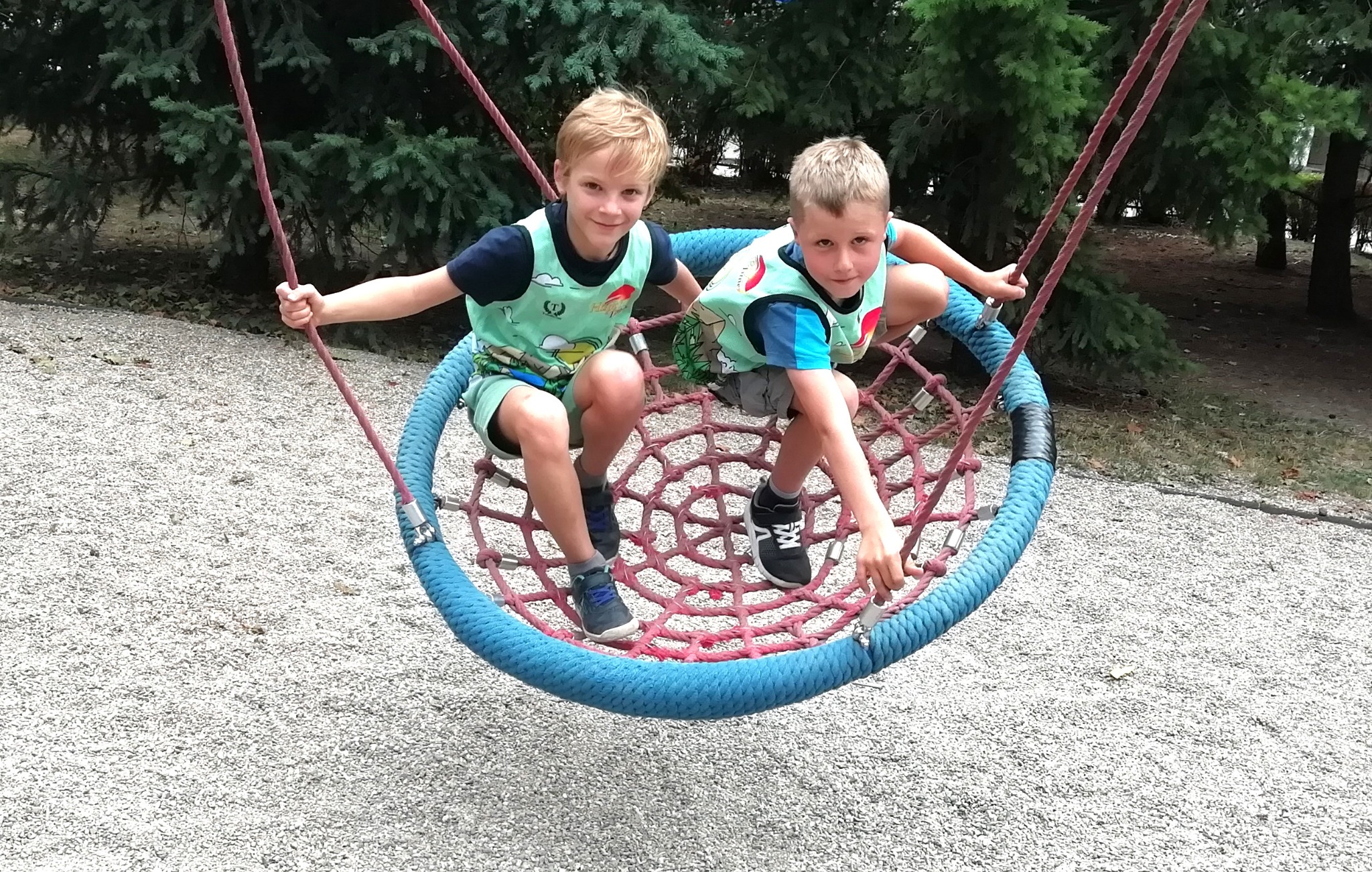 Dwóch chłopców w identycznych zielonych koszulkach siedzi na okrągłej huśtawce z siatki na placu zabaw.