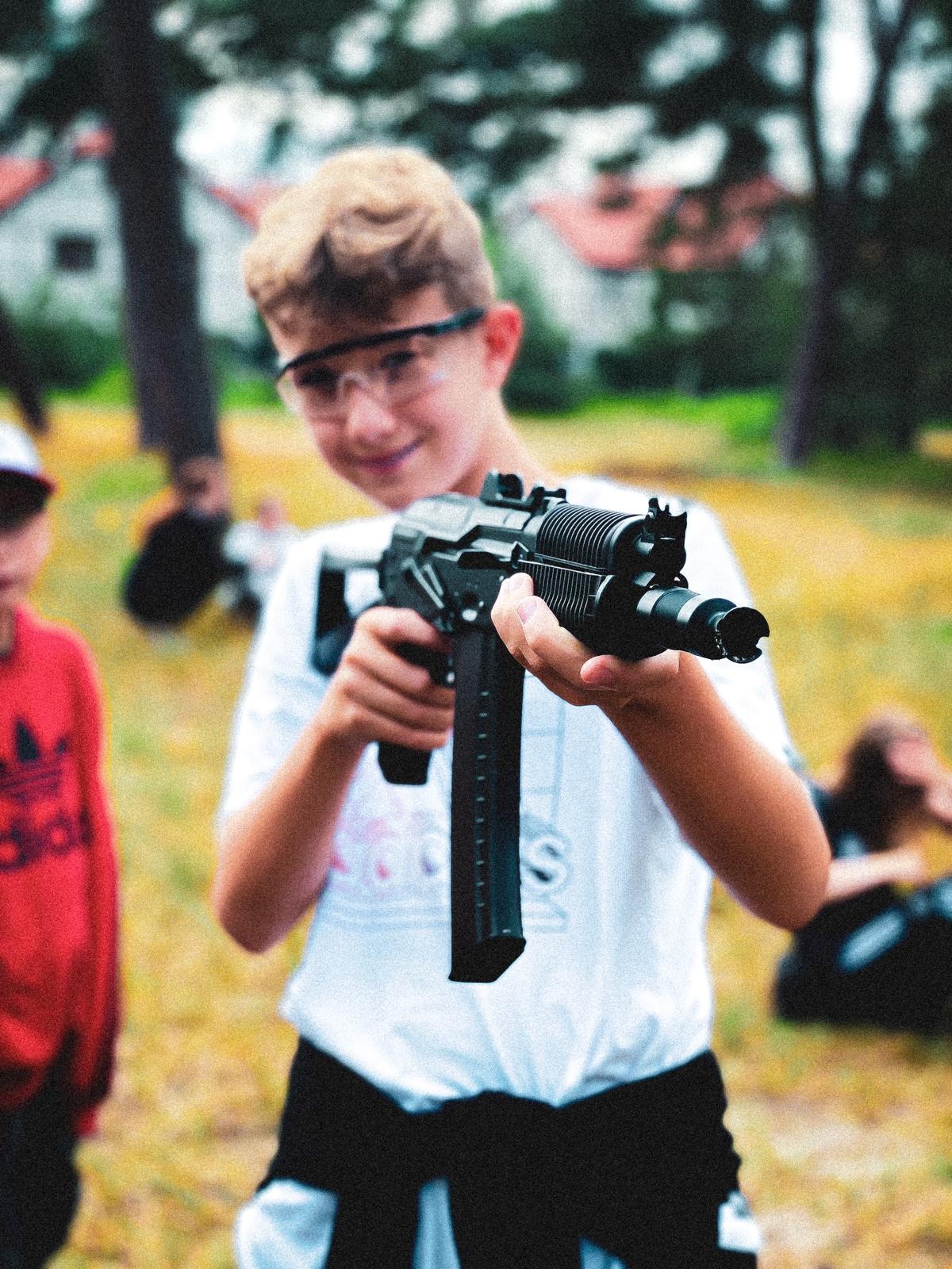 Uśmiechnięty nastolatek w okularach ochronnych celuje z repliki karabinu AK w plenerze, widoczni inni uczestnicy zabawy.
