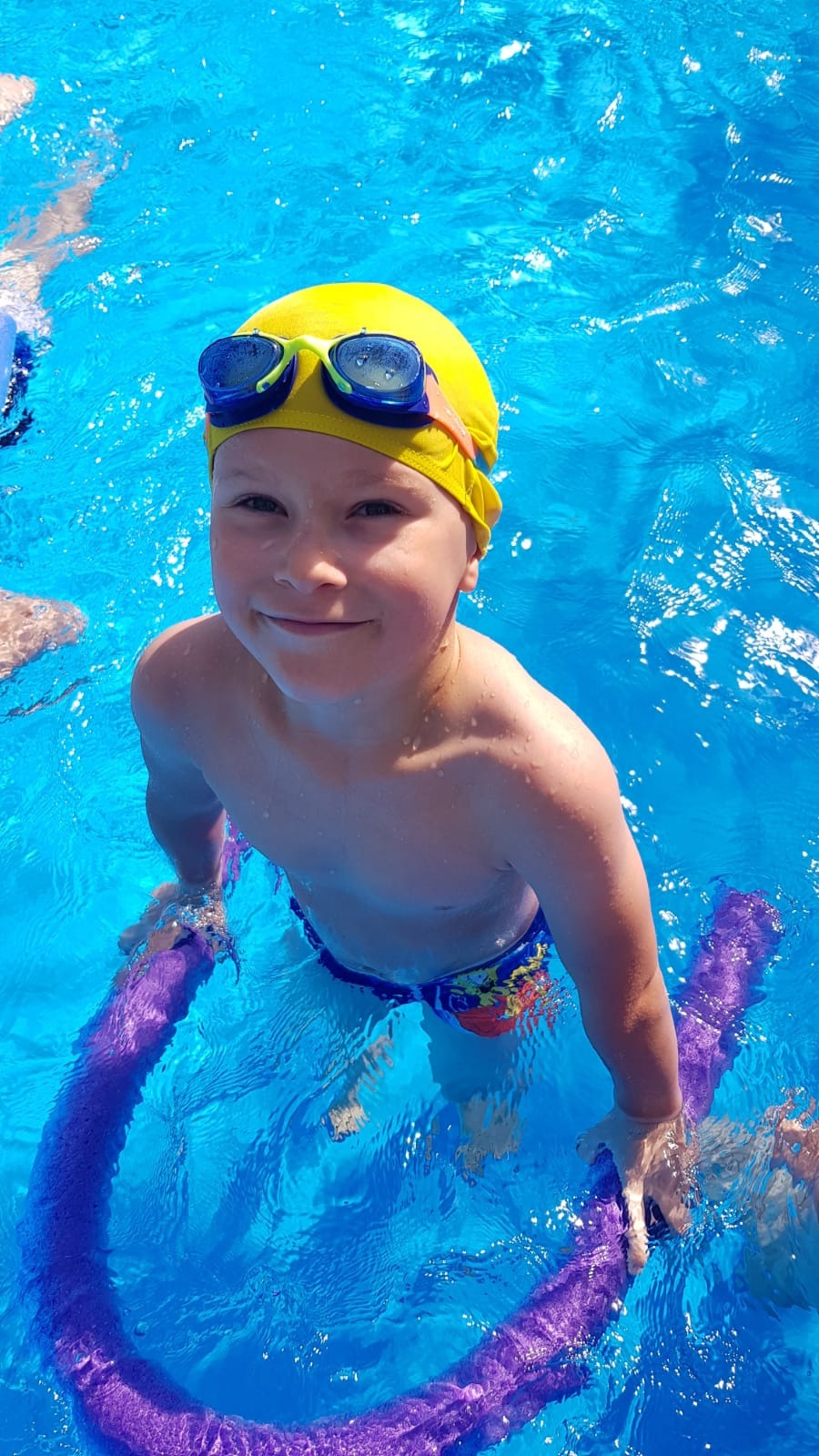 Uśmiechnięty chłopiec w żółtym czepku i niebieskich goglach pływa w basenie, trzymając fioletowy makaron piankowy.
