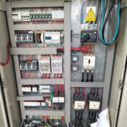 Wnętrze szafy sterowniczej z licznymi przewodami, przekaźnikami, bezpiecznikami i modułami sterującymi. Widoczne oznaczenia ostrzegawcze o napięciu. Przewody ułożone w wiązki, ale ogólny wygląd...