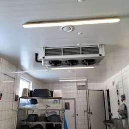 Agregat chłodniczy zamontowany pod sufitem w pomieszczeniu gospodarczym z metalowymi regałami i białymi kafelkami na ścianach.