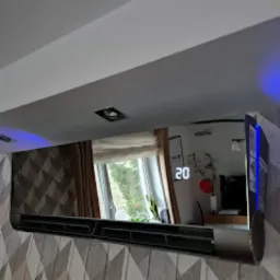 Czarny, lustrzany klimatyzator ścienny z widocznym wyświetlaczem temperatury (20 stopni), zamontowany w nowoczesnym wnętrzu z geometryczną tapetą i oświetleniem sufitowym LED.